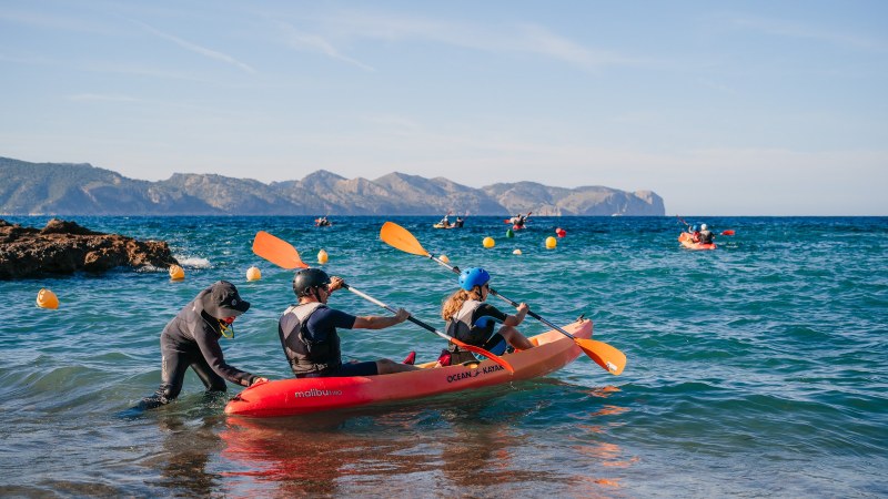 Person pushing kayak with two people into blue sea, mountains in the background.