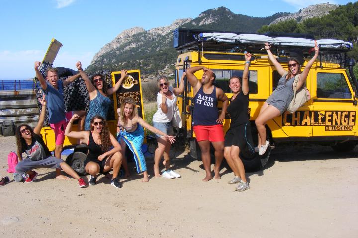 Group of people posing energetically in front of a yellow adventure vehicle with mountains in the background.