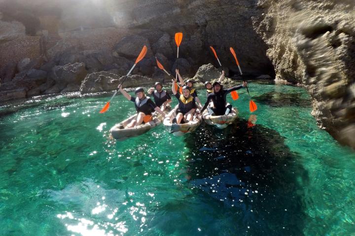 Group of people kayaking in clear turquoise water near rocky cliffs, holding paddles up.