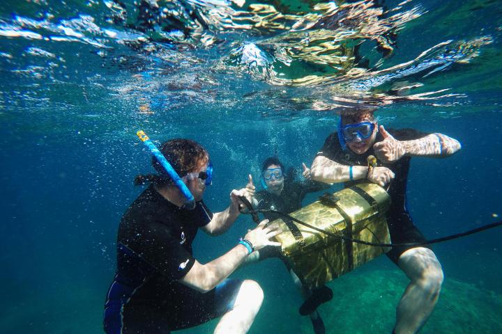 Three snorkelers underwater with a gold-colored chest, giving thumbs up.
