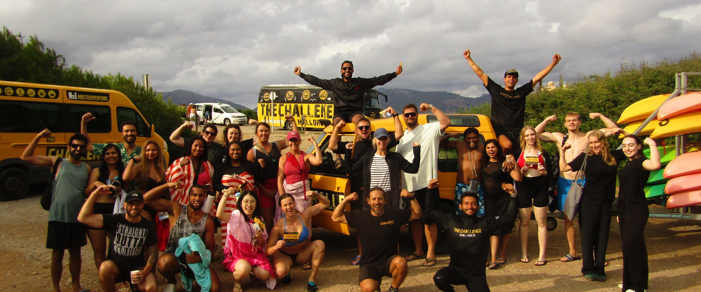 Group of people posing energetically in front of yellow vans and kayaks under a cloudy sky.