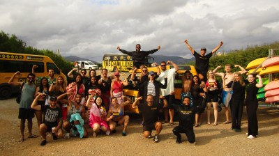 Group of people posing energetically in front of yellow vans and kayaks under a cloudy sky.