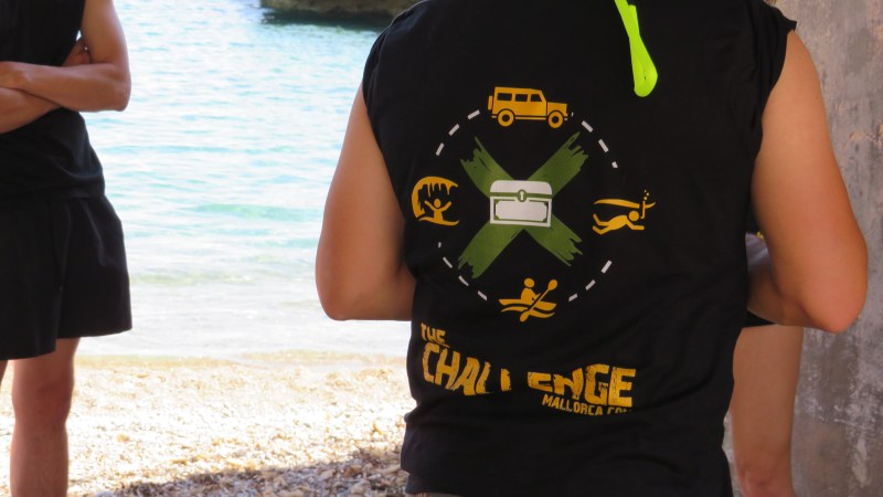 Person wearing 'The Challenge' shirt with adventure icons near beach.