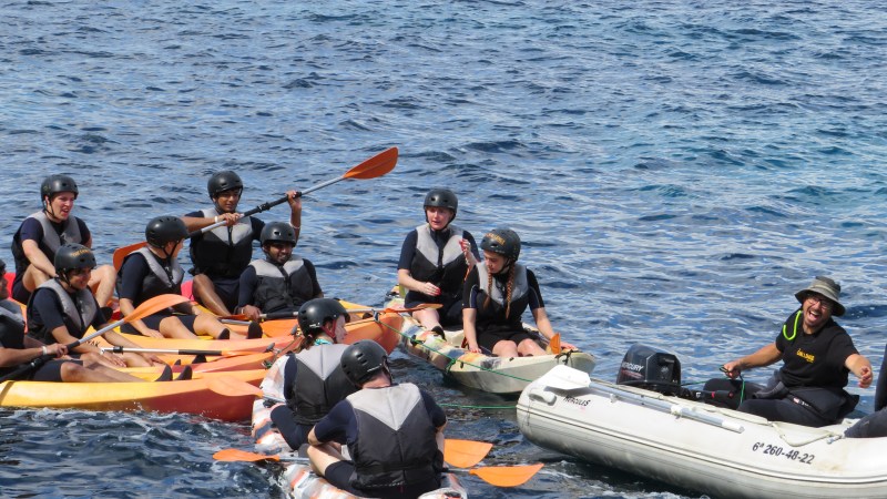 Group in kayaks and small boat on water, wearing helmets.