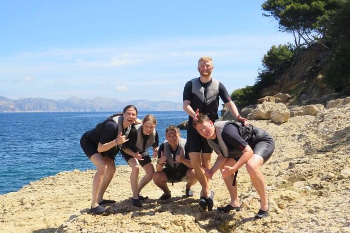 Group of people wearing wetsuits posing on a rocky beach by the sea.