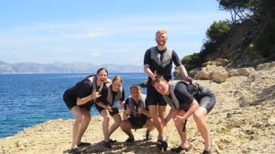 Five people in wetsuits posing on rocky shore with ocean background.