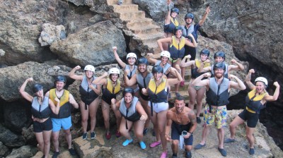 Group of people in helmets and life jackets posing on rocky steps with raised fists.