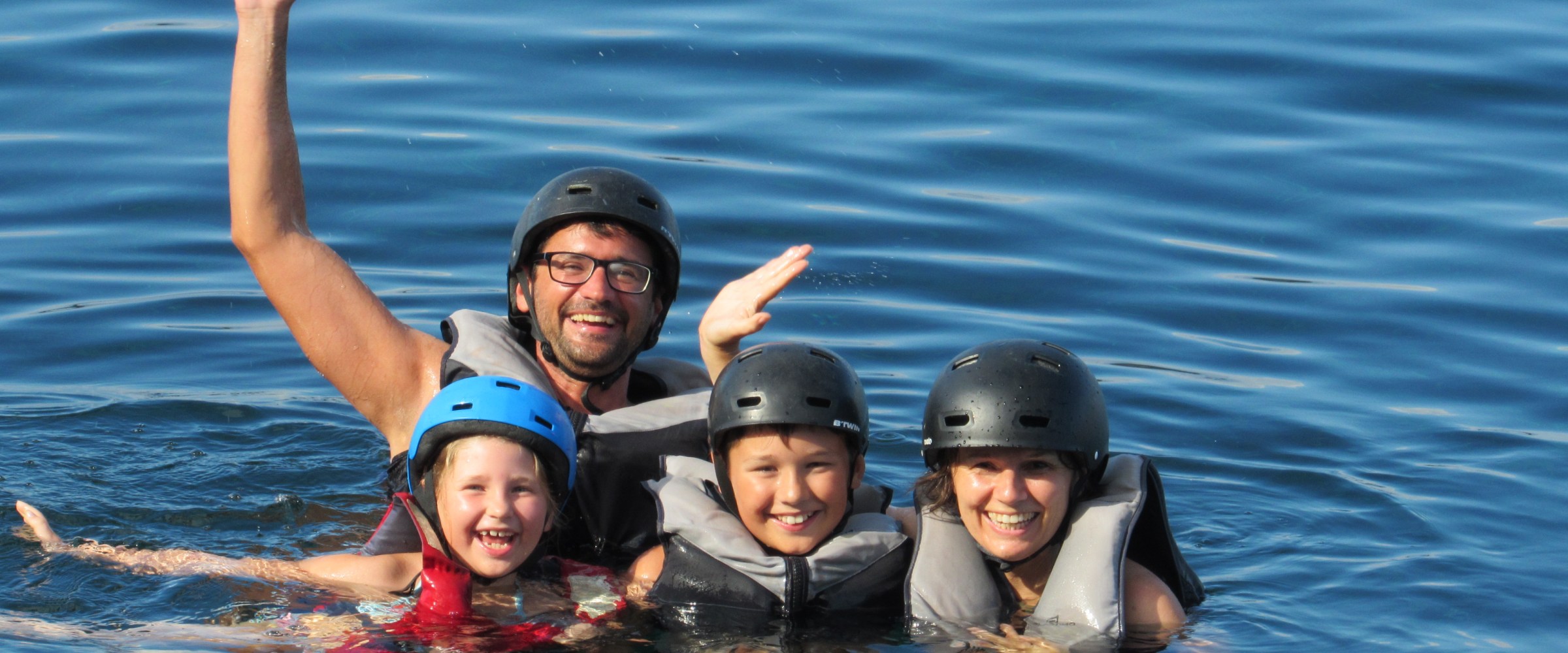 Four people wearing helmets and life jackets smiling and waving while swimming in a lake.