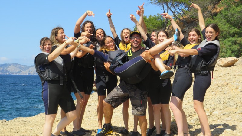 Group of people in wetsuits cheerfully posing on a rocky shoreline, one person being lifted in the air.