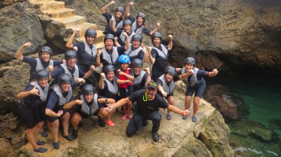Group of people in helmets and wetsuits posing energetically on rocky outcrop near the water.