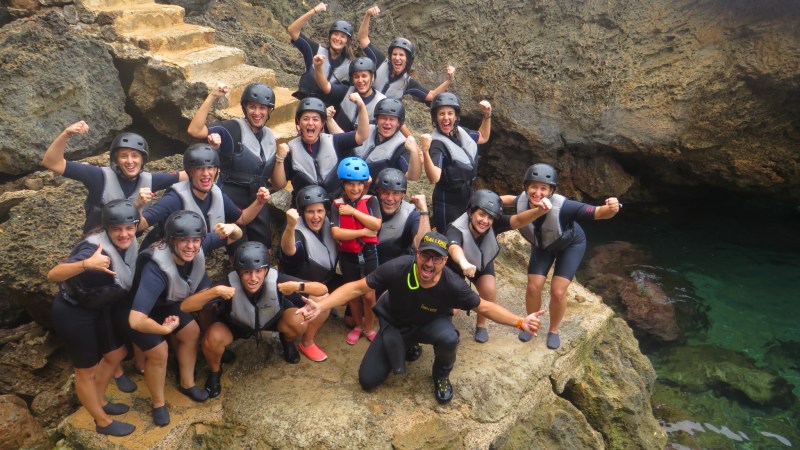 Group of people in helmets and wetsuits posing energetically on rocky outcrop near the water.