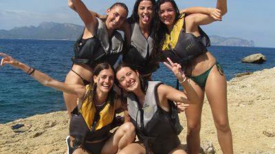 Five women in life vests posing on a rocky shore with the sea in the background.