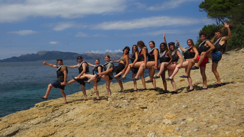 Group of people in life vests posing with legs raised on rocky shore.