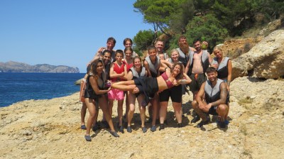 Group of people in swimwear posing on rocky beach with ocean and trees in the background.