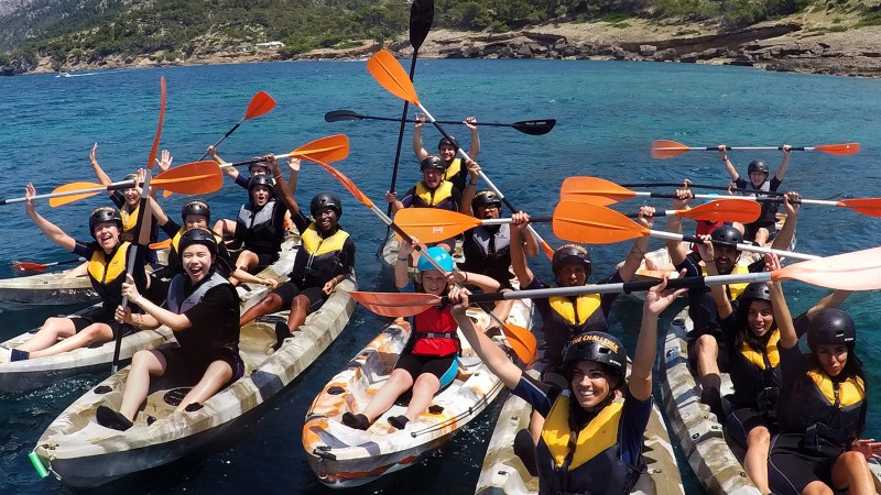Group of kayakers on the sea with paddles raised, near rocky shore.