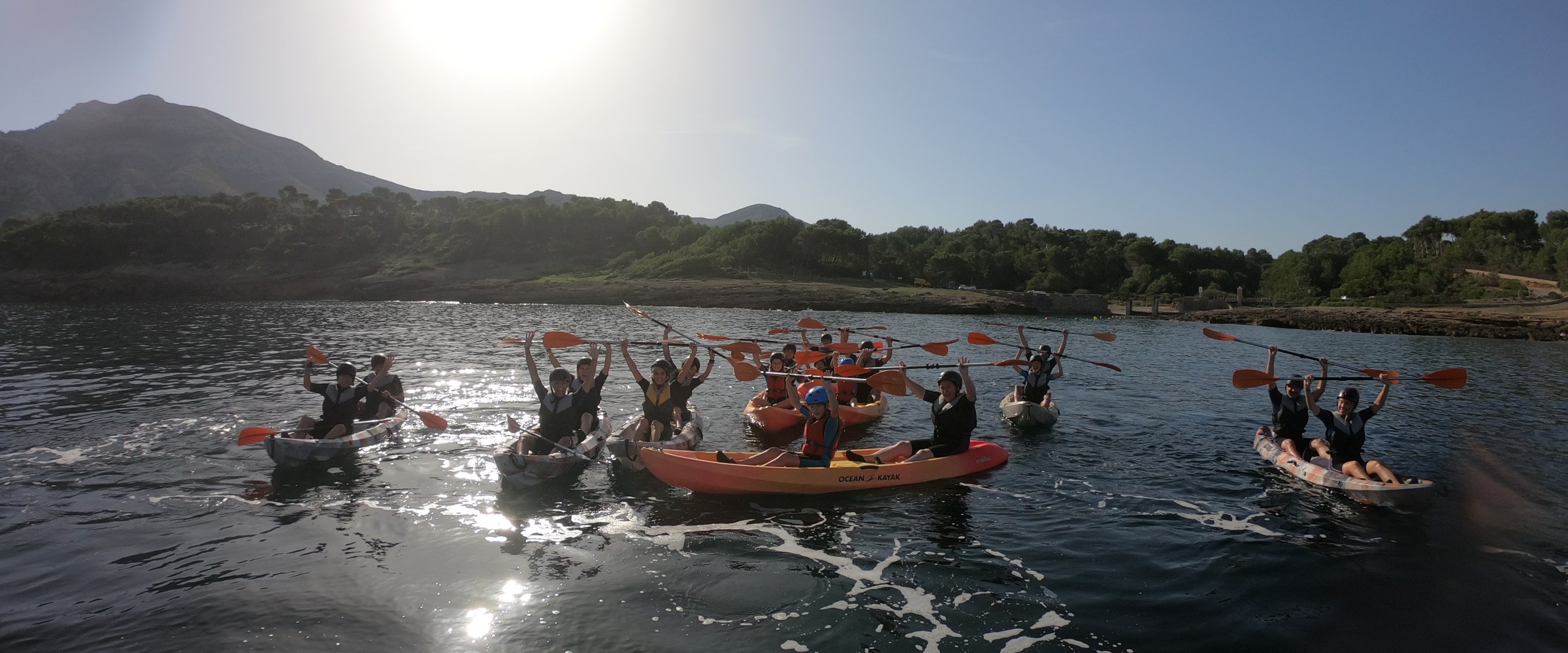 Group kayaking on calm water under bright sun with distant hills.