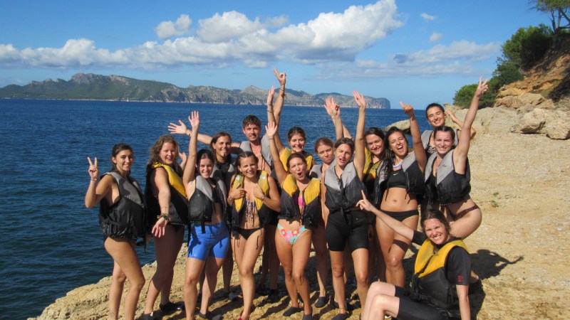 Group of smiling people in life vests posing on rocky shore by the sea.