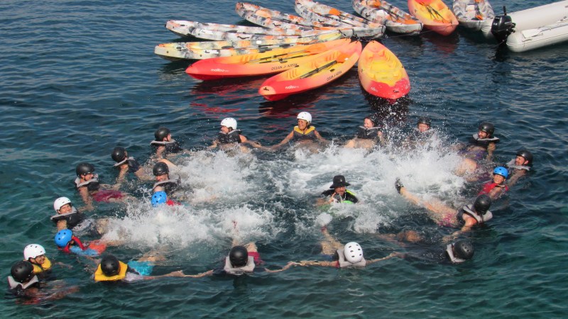 People in helmets hold hands in a circle, splashing water, with kayaks floating nearby.